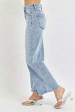 A female posing from the side wearing light blue RISEN Full Size Tummy Control High Rise Crop Wide Leg Jeans paired with a floral white blouse, jewelry, and white round open-toe heels.