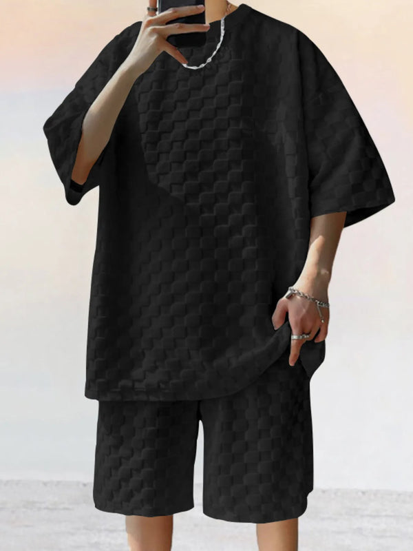 Front view of man taking a selfie photo wearing black Therion Boldline Set with round neck checkered shirt and shorts, styled with sneakers and accessories.