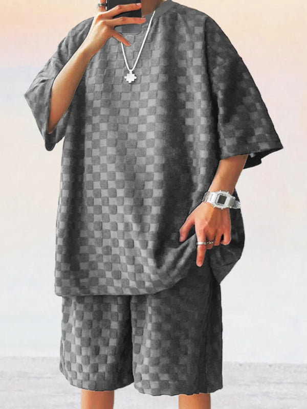 Front view of man taking a selfie photo wearing dark gray Therion Boldline Set with round neck checkered shirt and shorts, styled with sneakers and accessories.