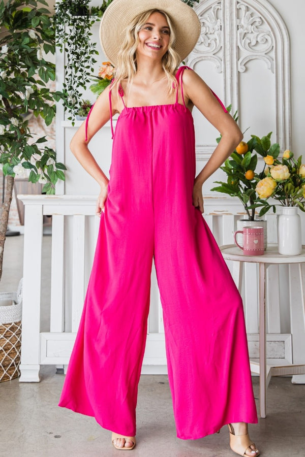 Model posing in a garden wearing a Fuchsia Veveret Pocketed Spaghetti Strap Wide Leg Jumpsuit paired with a straw hat and tan heels.