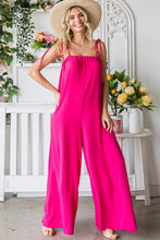 Full front view of the model in a Fuchsia Veveret Pocketed Spaghetti Strap Wide Leg Jumpsuit styled with a straw hat and tan heels in a garden setting.