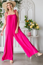 Side angle of a model posing in a garden wearing a Fuchsia Veveret Pocketed Spaghetti Strap Wide Leg Jumpsuit with a straw hat and tan heels.