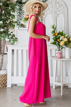 Model strolling through a garden in a Fuchsia Veveret Pocketed Spaghetti Strap Wide Leg Jumpsuit styled with a straw hat and tan heels.