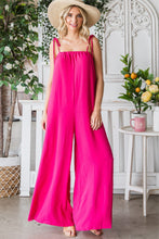 Model walking through a garden setting in a Fuchsia Veveret Pocketed Spaghetti Strap Wide Leg Jumpsuit paired with a straw hat and tan heels.