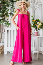 Model seated in a garden wearing a Fuchsia Veveret Pocketed Spaghetti Strap Wide Leg Jumpsuit styled with a straw hat and tan heels.