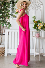 Side angle of a model posing in a garden wearing a Fuchsia Veveret Pocketed Spaghetti Strap Wide Leg Jumpsuit with a straw hat and tan heels.