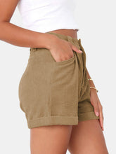 Close up photo of a model highlighting the pocket detailing of the Camel Metropolitan Maven High-Waist Shorts from the side paired with a white tank top and jewelry.