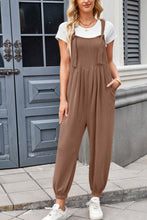 Model wearing brown Xévia Knotted Strap Overalls with a fitted tank top and sneakers, standing against a neutral wall.