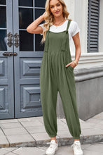Model styling dark green Xévia Overalls with a white t-shirt, layered necklaces, gold hoop earrings, a bangle, and casual white sneakers for an easygoing daytime look.