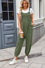 Model casually leaning against a wall in dark green Xévia Overalls paired with a white tee, gold jewelry, and white sneakers, holding a coffee cup.