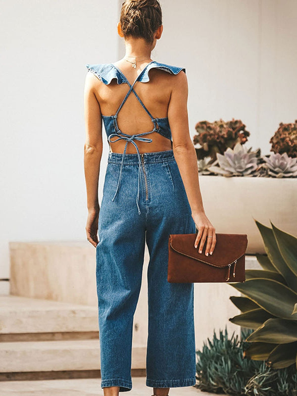 Model showing the back of the Zelisse Denim Jumpsuit in medium blue, highlighting the backless criss-cross adjustable straps, tie detail, and back zipper closure, styled with a brown clutch and pendant necklace.