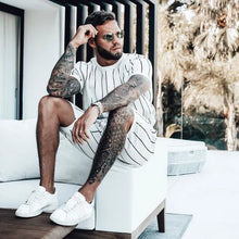 Man wearing white lounge set with black stripes, legs propped up, showcasing shorts and casual styling.